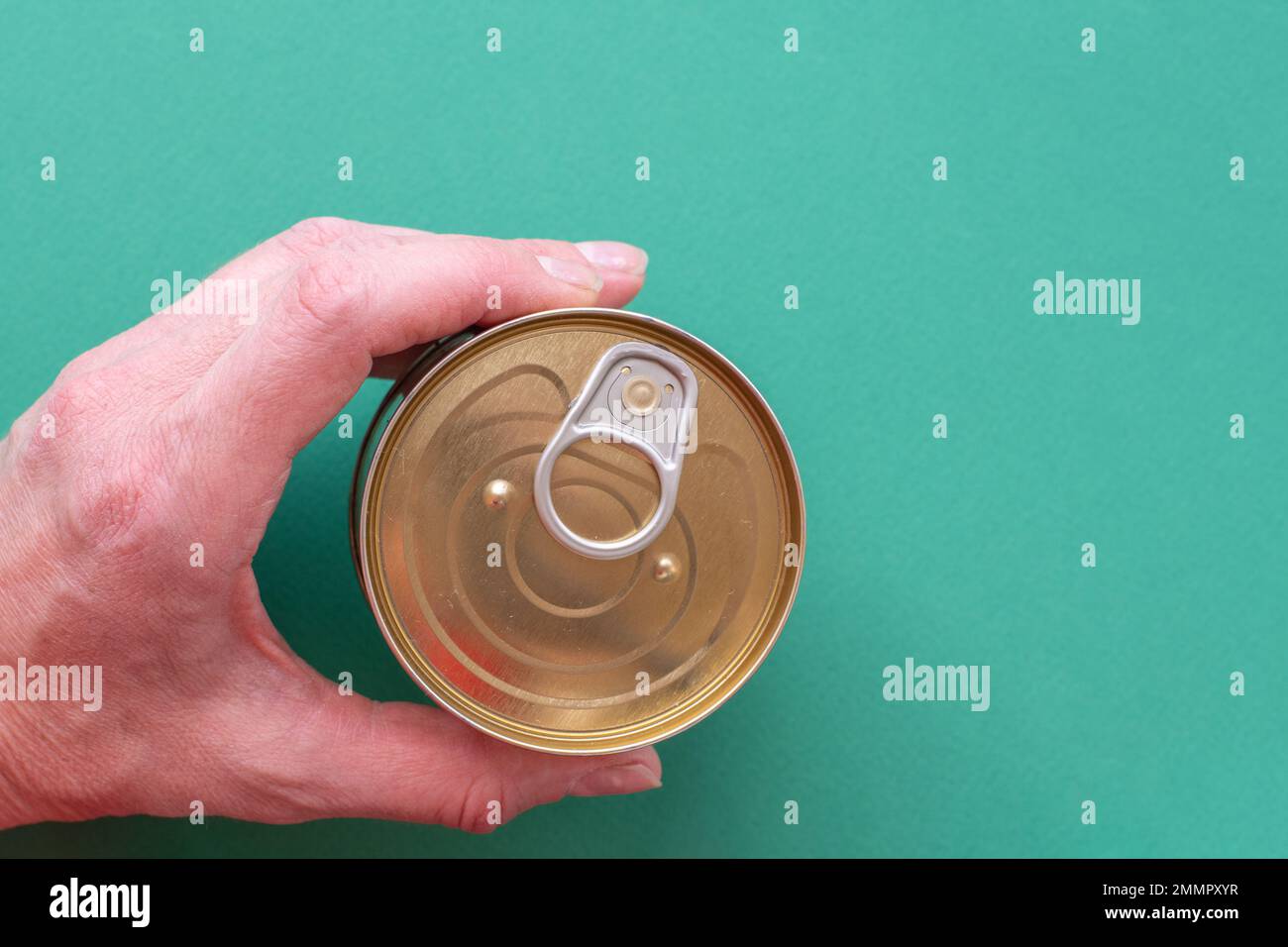 The hand of an adult man holds a closed can of canned food with a public key on a green background. Top view of tin can with ring pull isolated on gre Stock Photo