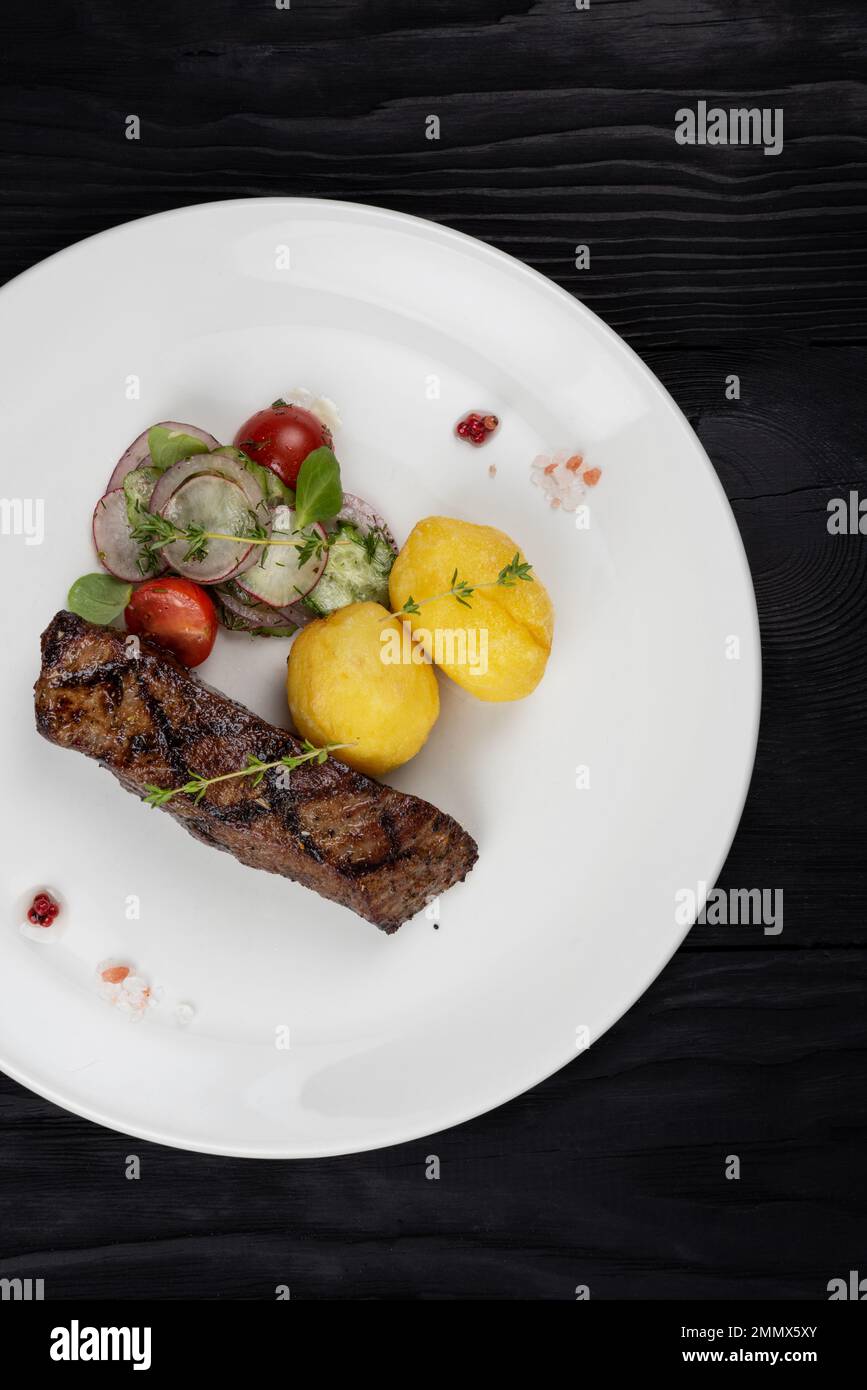 Grilled beef skirt steak meat with potato and vegetables on white plate on wooden black background Stock Photo