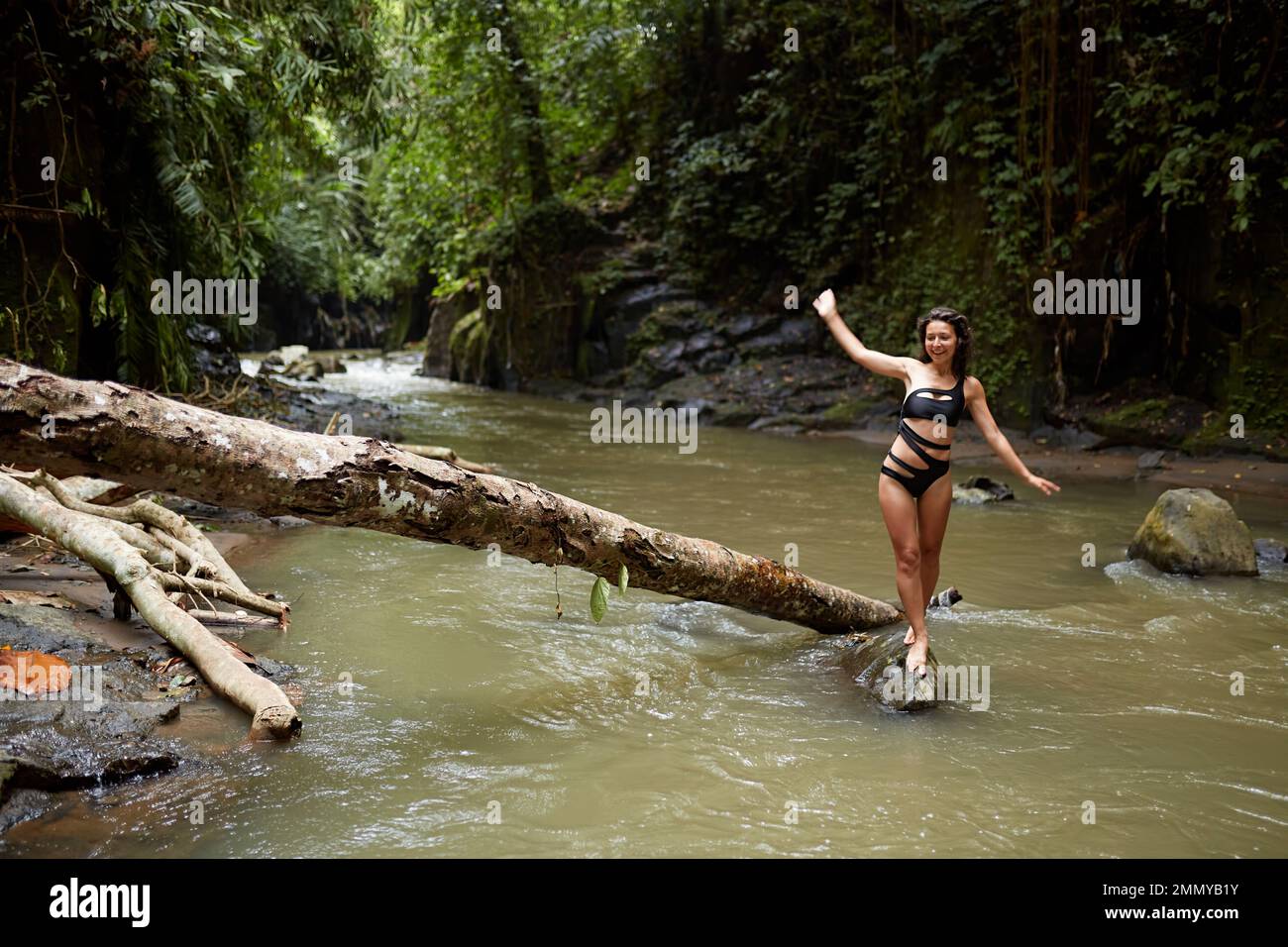 A young, slender girl poses on a tree trunk that has fallen into the river in the middle of a dense jungle. Stock Photo