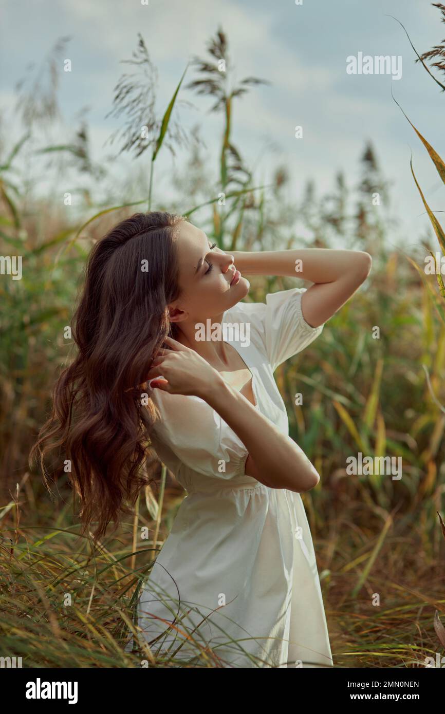 Beautiful young woman in white summer dress is sitting in tall grass in rural field. Portrait of romantic girl at sunset, warm sun, natural beauty wom Stock Photo