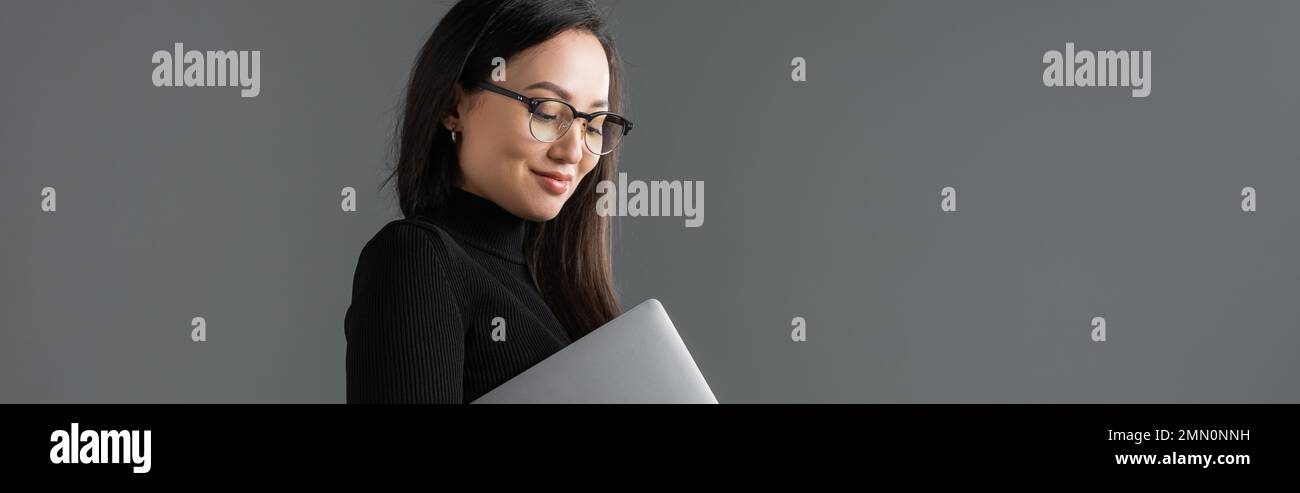 cheerful asian woman in black turtleneck and glasses holding laptop ...
