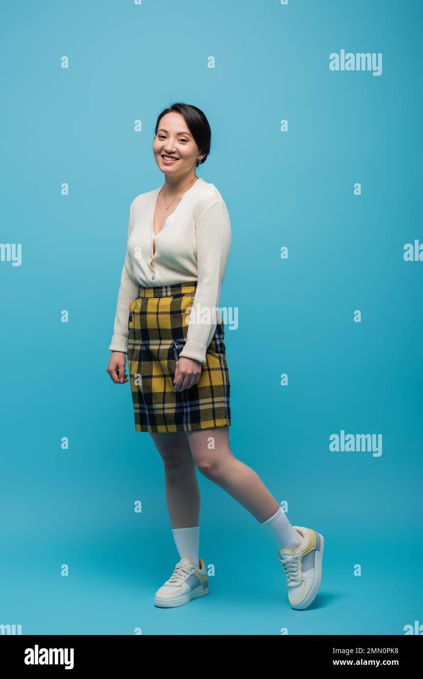 full length of cheerful asian woman in checkered skirt and sneakers