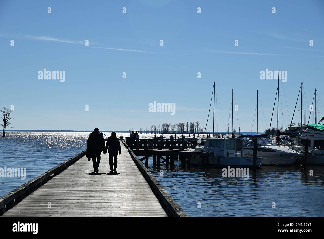 A couple holds hands is silhouetted while walking down a wooden boat dock. Stock Photo