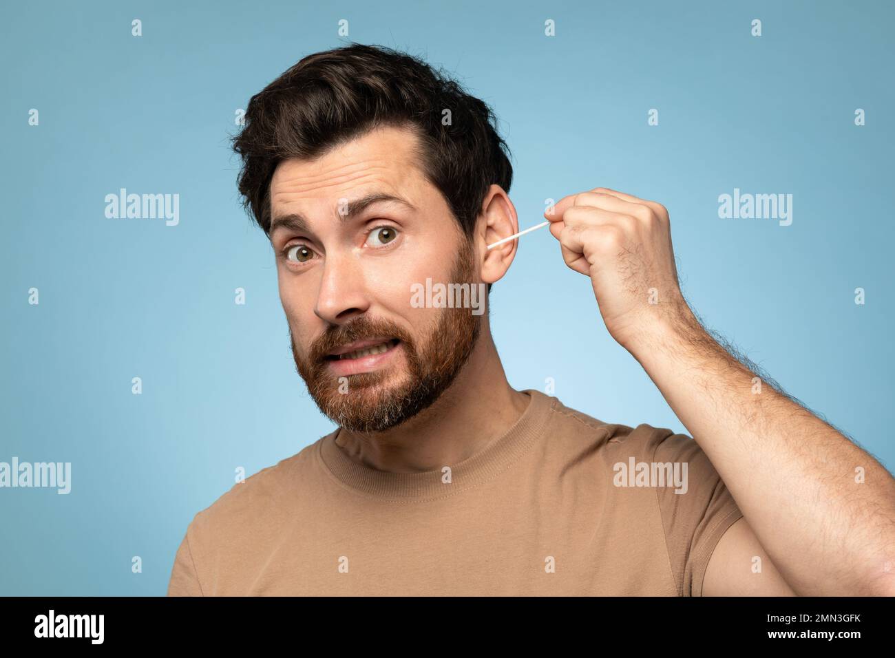 Funny middle aged bearded man cleaning ears with cotton swab, doing daily hygiene routine, blue studio background Stock Photo