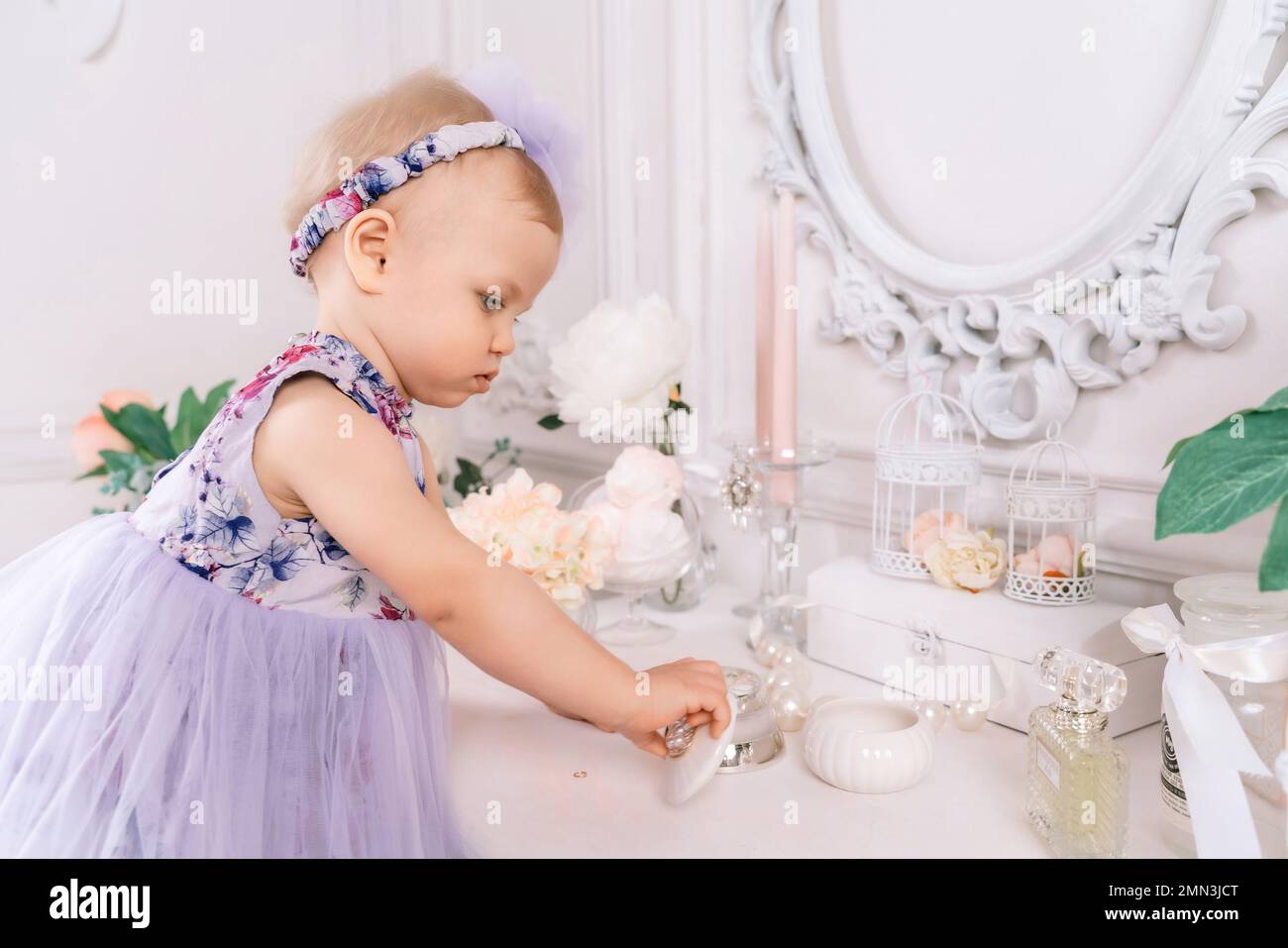 Baby girl elegant dress. A one-year-old girl in a puffy dress and a cute bow poses against the backdrop of a bright room with a dressing table and Stock Photo