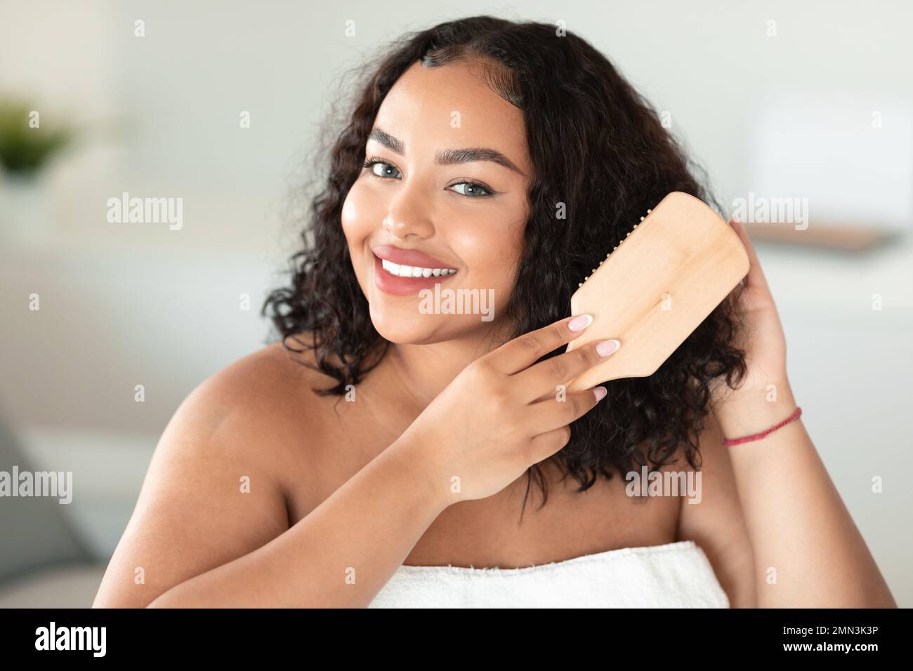 Portrait of happy body positive lady brushing her curly dark hair and smiling at camera, home interior Stock Photo