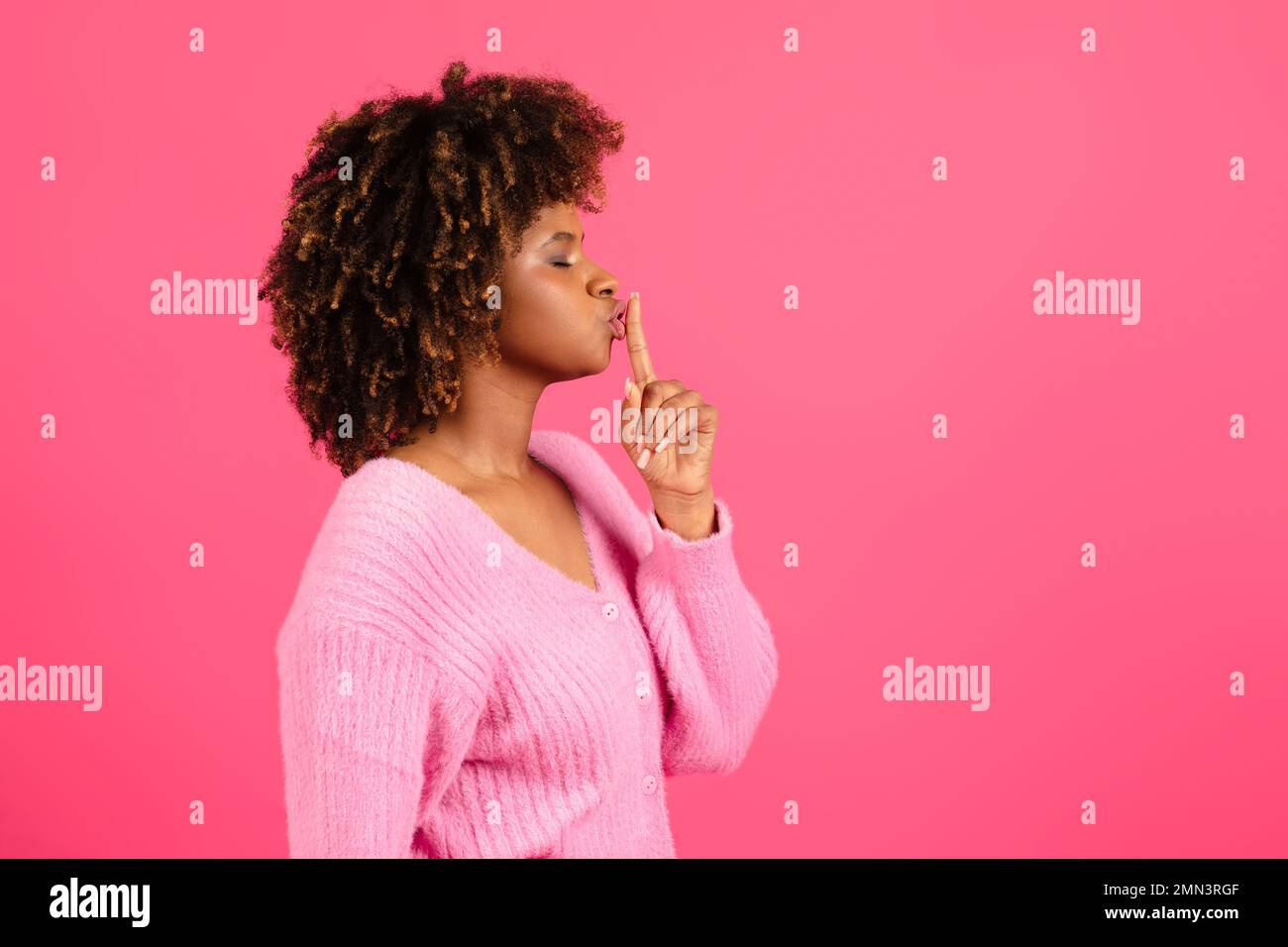 Serious young black curly female in casual presses her finger to lips and makes shhh sign Stock Photo