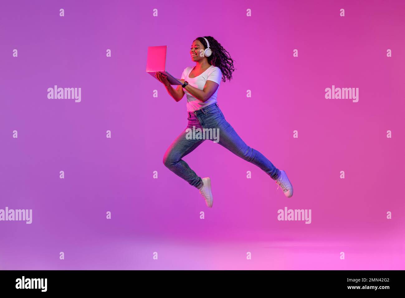 Happy Black Woman In Wireless Headphones Jumping With Laptop In Neon Light Stock Photo
