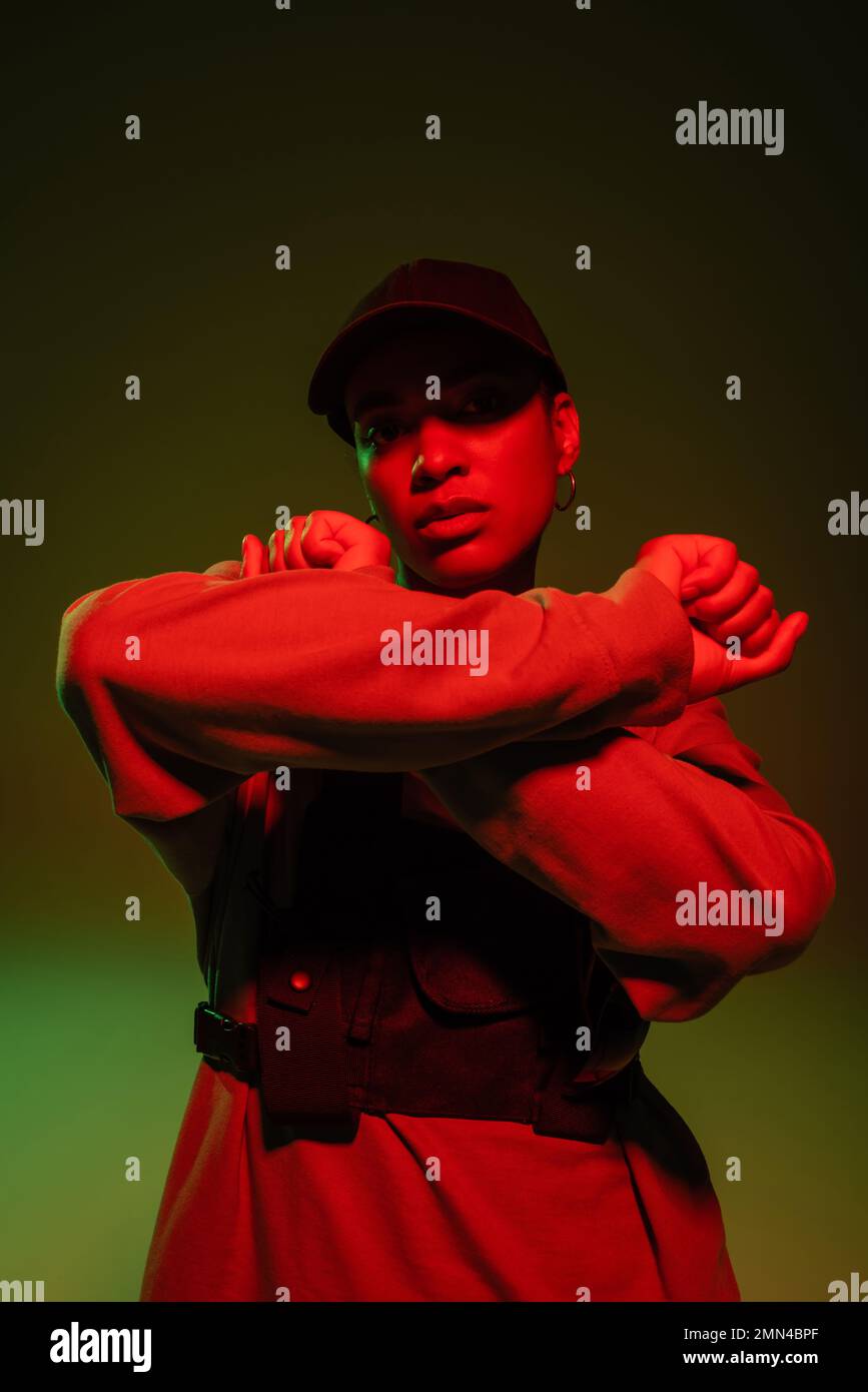 african american woman in sweatshirt and baseball cap posing with crossed arms on green with red light Stock Photo