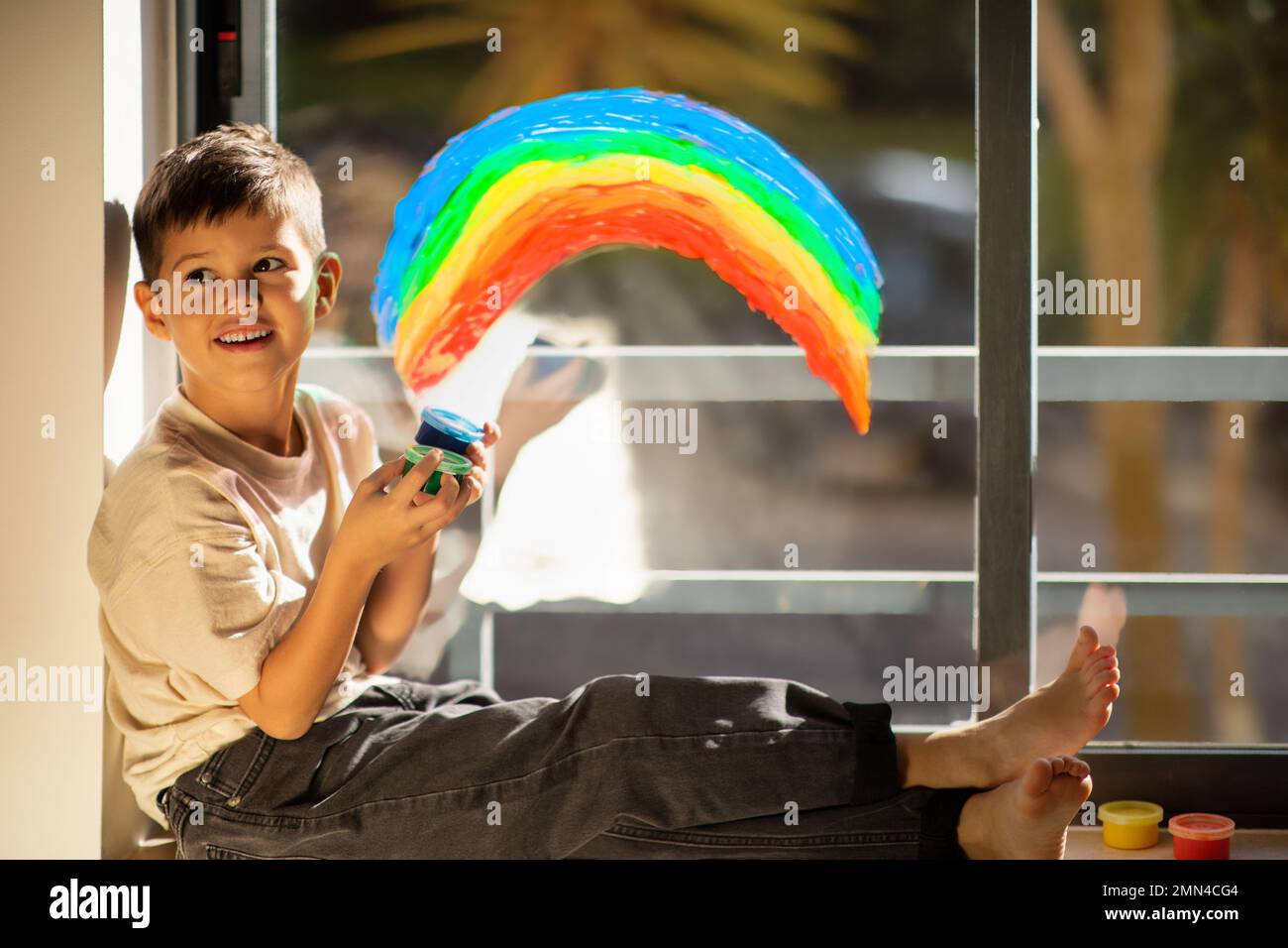 Happy small european boy chooses paints, draws rainbow on window in children room interior Stock Photo