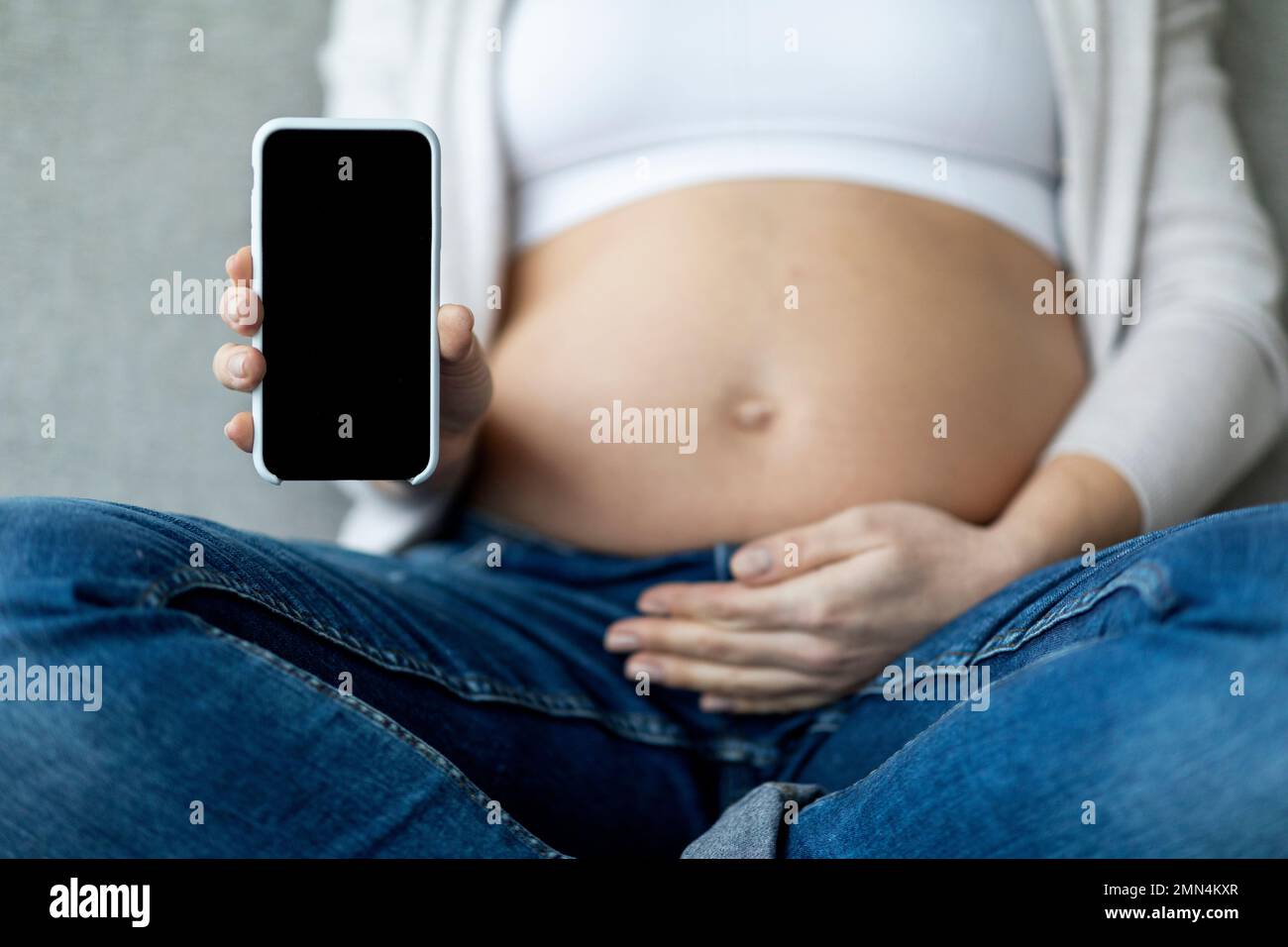 Unrecognizable Pregnant Woman Showing Blank Smartphone And Embracing Belly At Home Stock Photo