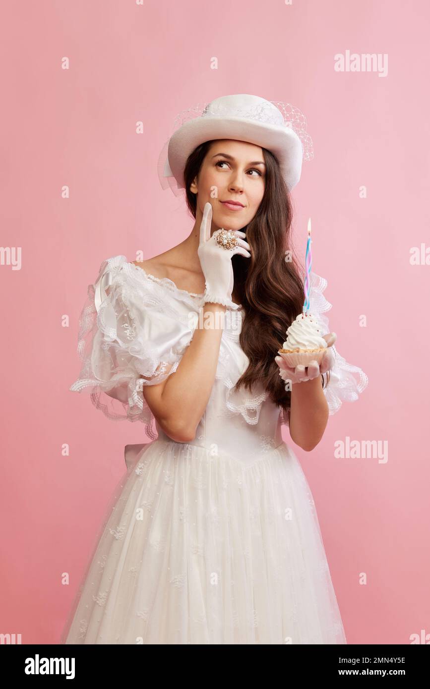 Making wishes. Portrait of beautiful lady in white vintage dress posing with birthday cupcake over pink background. Concept of 19th century, fashion Stock Photo