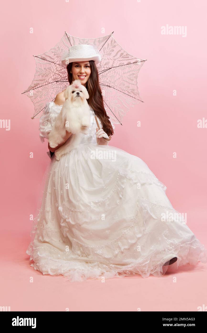 Portrait of beautiful lady, woman in white vintage dress posing with cute dog over pink background. Concept of 19th century, fashion, comparison of Stock Photo