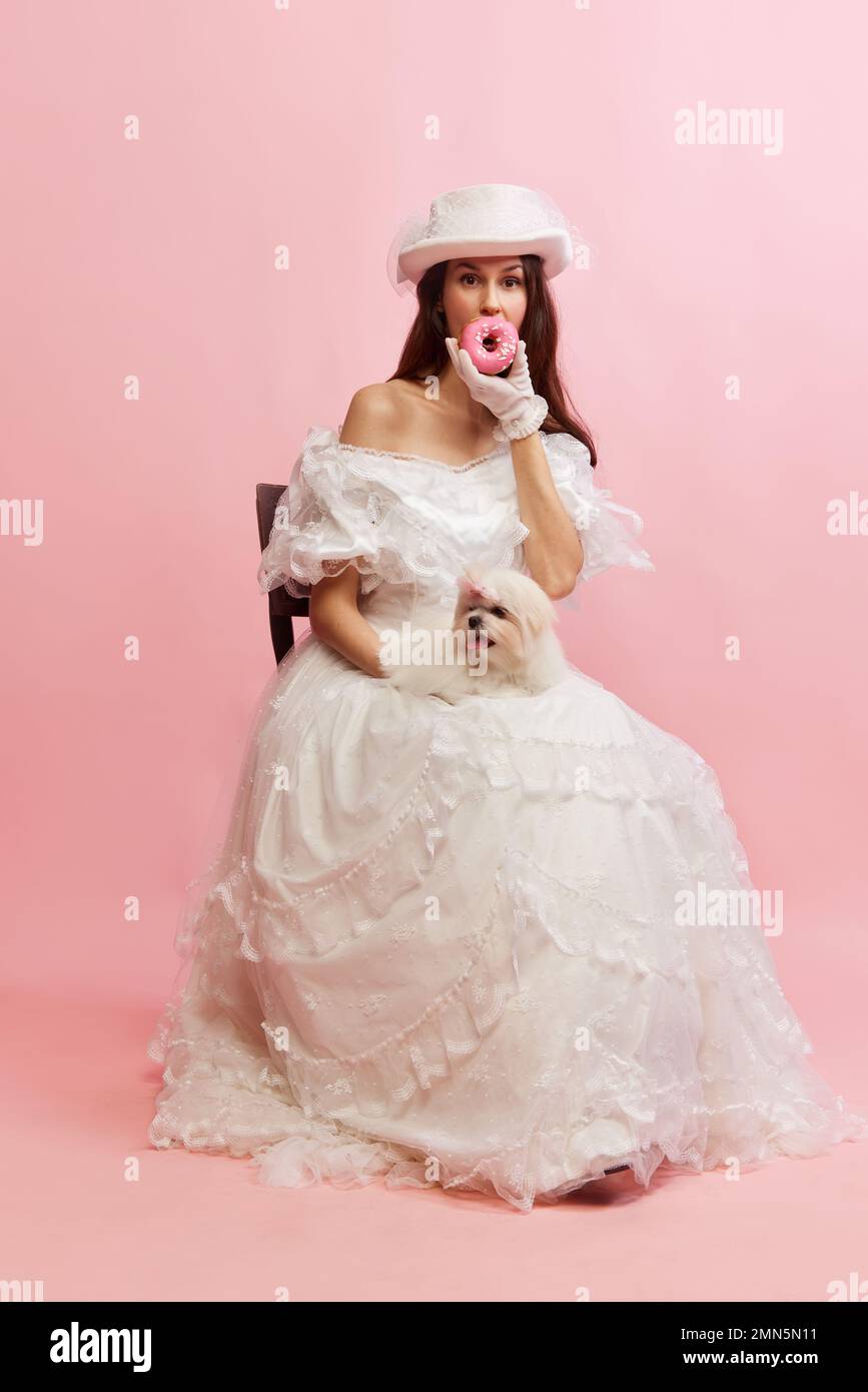 Eating donuts. Portrait of beautiful lady in white vintage dress posing with little dog over pink background. Concept of 19th century, fashion Stock Photo