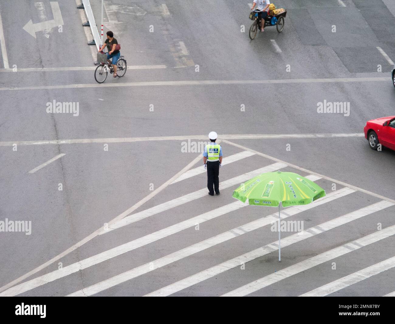 Somewhat isolated and perhaps lonely traffic officer policeman at a road junction in the Chinese city of Xi'an. PRC. China. Guiding and directing a small amount of traffic but looking exposed. He has a large parasol umbrella for shade should he need it. (125) Stock Photo