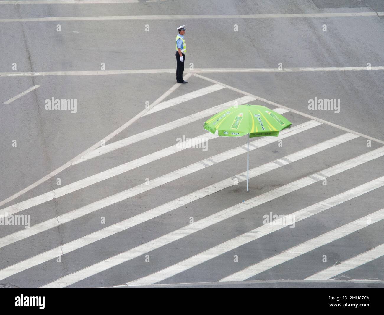 Somewhat isolated and perhaps lonely traffic officer policeman at a road junction in the Chinese city of Xi'an. PRC. China. Guiding and directing a small amount of traffic but looking exposed. He has a large parasol umbrella for shade should he need it. (125) Stock Photo
