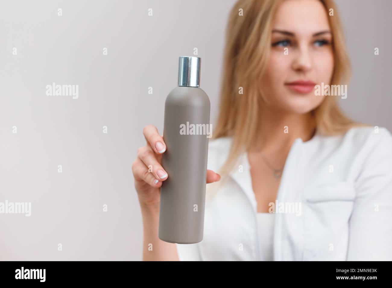 Blonde nurse in white coat hold blank plastic bottle with cosmetics on plain background Stock Photo