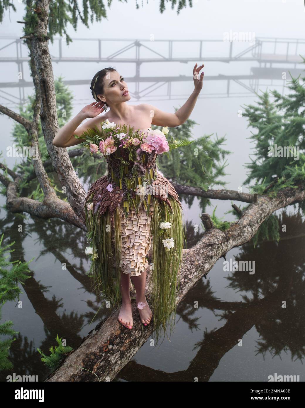 Blooming gorgeous lady in dress of flowers on tree over water in the morning fog Stock Photo
