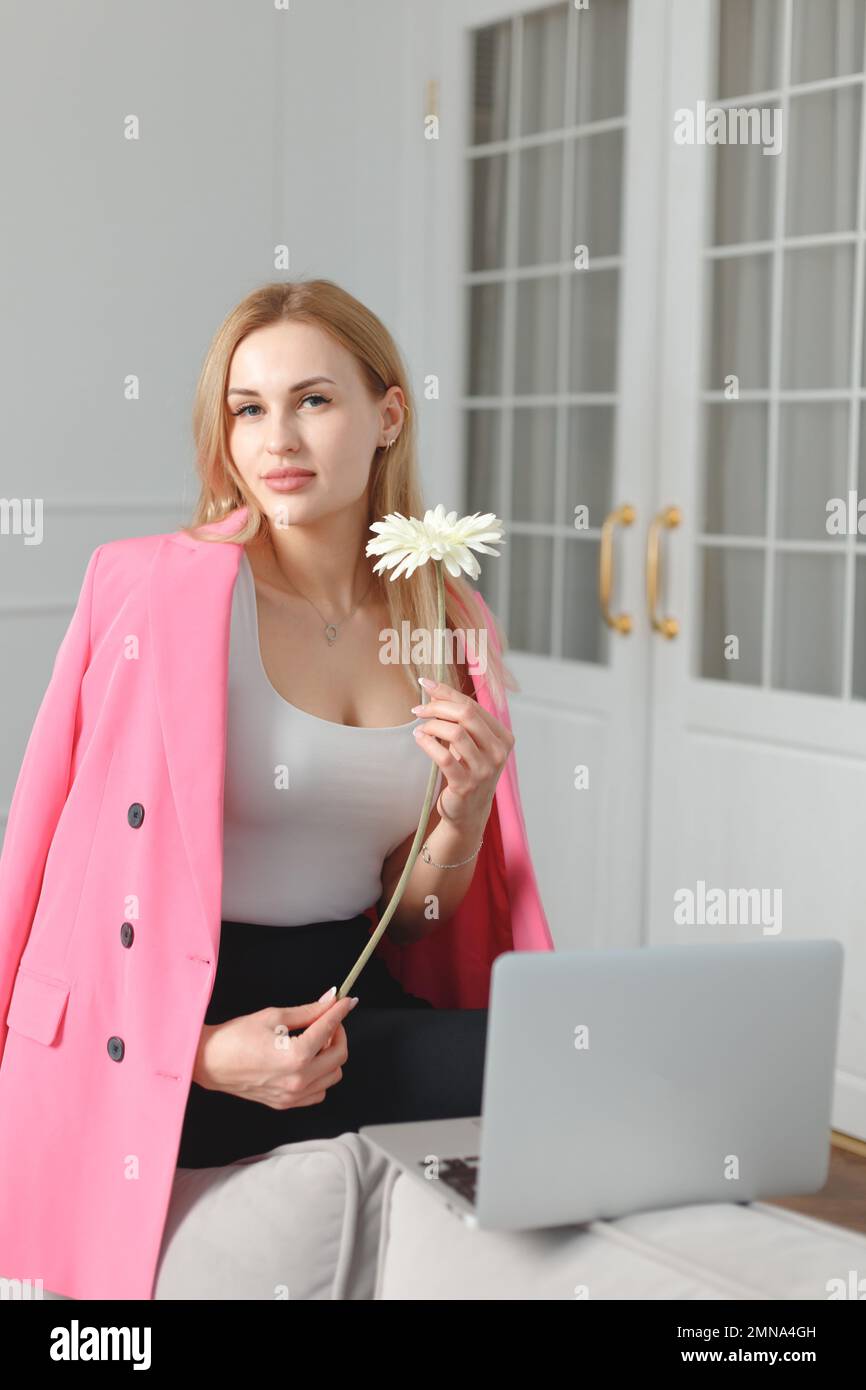 Blonde young woman in pink blazer with laptop and flower working in living room on coach at home. Female feeling, harmony, psychological balance Stock Photo