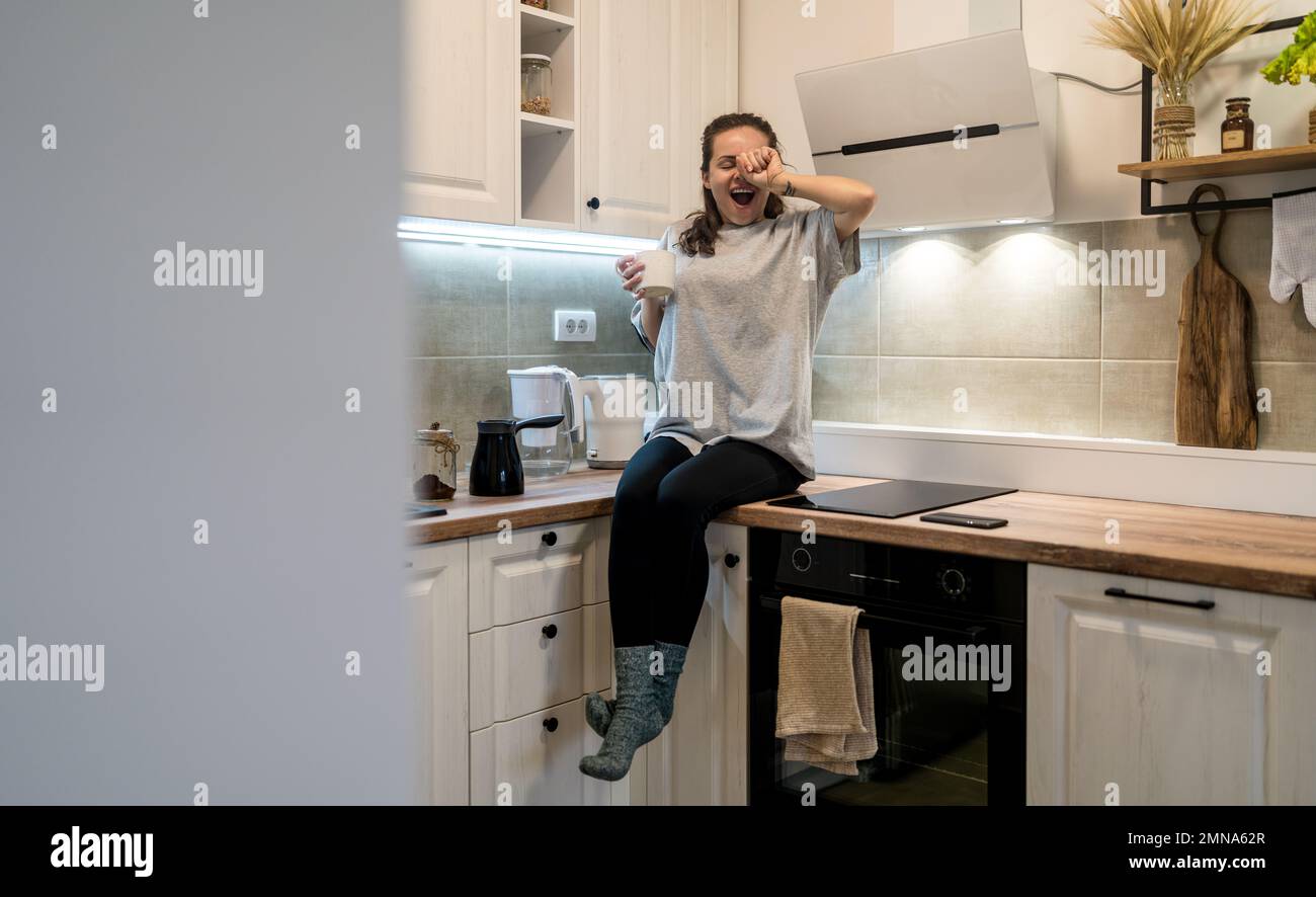 Woman sits on the kitchen furniture holding coffee mug in hand yawning and rubbing eye with her hand, morning life concept. Stock Photo