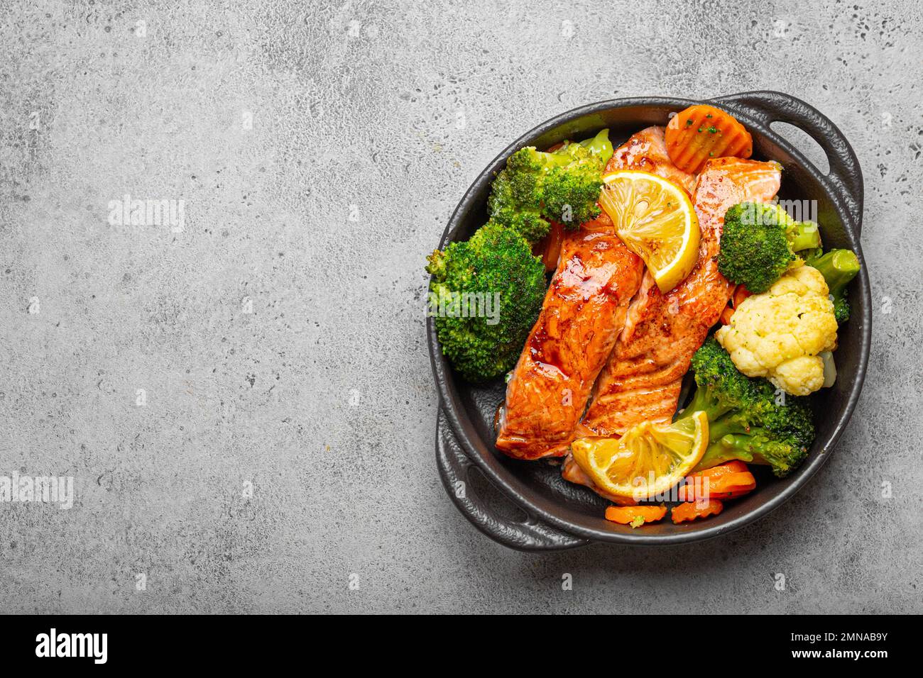 Healthy baked fish salmon steaks, broccoli, cauliflower, carrot in black cast iron casserole bowl on grey stone background. Cooking a delicious low Stock Photo