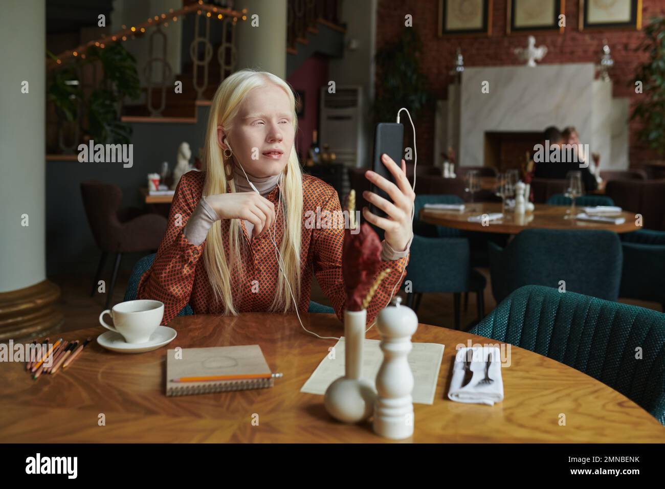 Albino girl with long hair looking at smartphone screen during communication with friend in video chat in cozy cafe Stock Photo