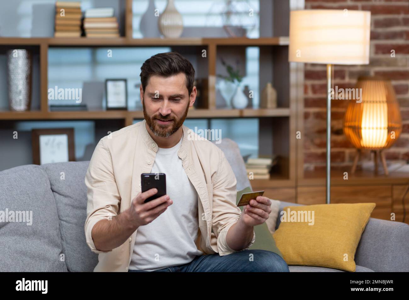 A young handsome man in a beige shirt is sitting on the sofa at home, using a phone and a credit card. Stock Photo