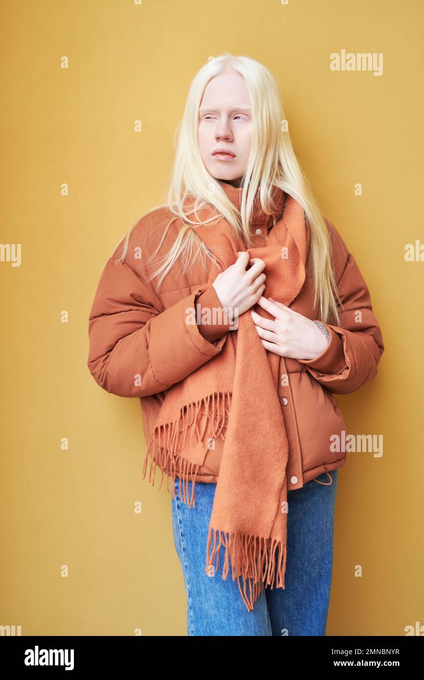 Long haired albino girl in blue jeans, brown jacket and scarf standing against yellow wall or background in isolation and looking aside Stock Photo