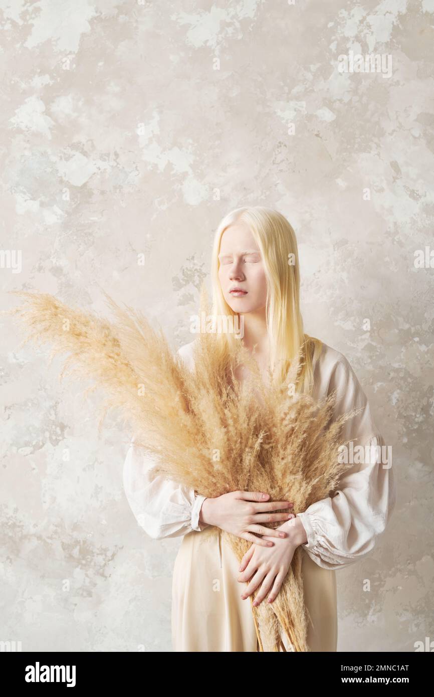 Young albino woman with bunch of fluffy dried flowers in hands keeping her eyes closed while standing by wall in front of camera Stock Photo