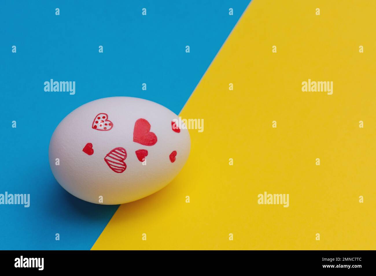 White Easter egg with various red hearts on yellow-blue background. Concept of Ukrainian refugees in search of safety and love for Ukraine. Copy Space Stock Photo