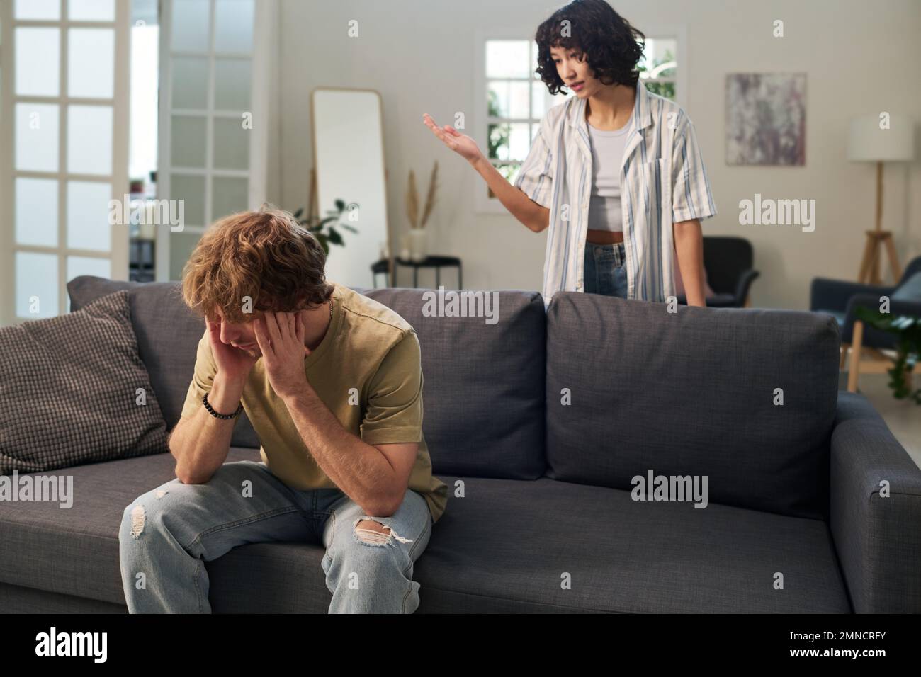 Young annoyed man sitting on sofa and touching his head while his irritated wife standing behind and scolding him during quarrel Stock Photo
