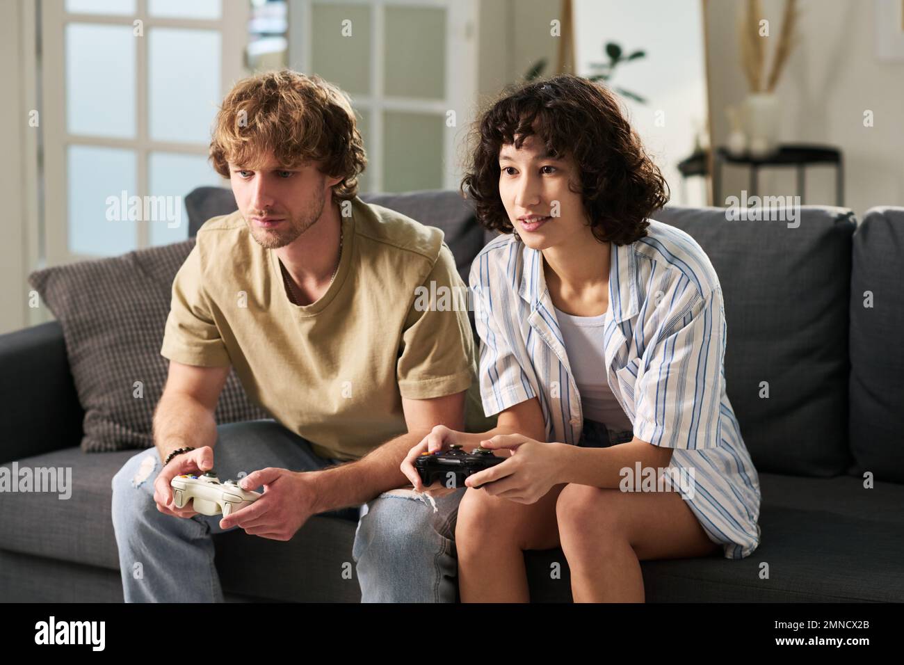 Young couple with control pads looking at screen of tv set while sitting on couch in living room at leisure and playing video games Stock Photo