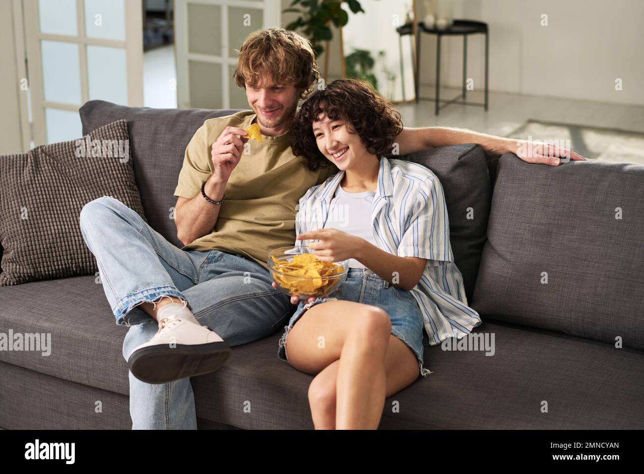 Happy young husband and wife laughing while sitting on couch in front of tv set, eating potato