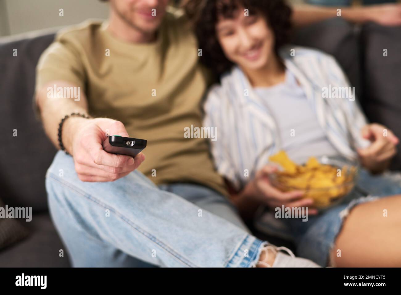 Focus on hand of young restful man holding remote control while sitting next to his wife with potato chips and choosing tv channel Stock Photo