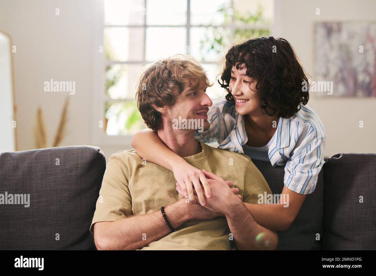 Happy young brunette woman embracing her husband sitting on couch and looking at him with smile while spending weekend at home Stock Photo