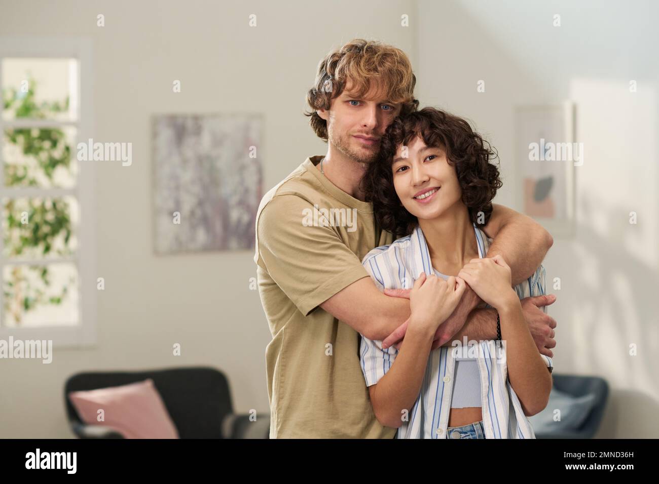 Happy young brunette woman in casualwear enjoying embrace of her husband while both standing in living room and looking at camera Stock Photo