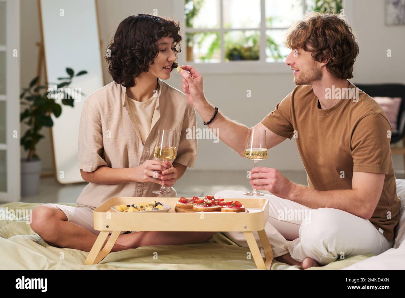 Happy young couple enjoying snack on tray and toasting with champagne while celebrating their marriage on bed in hotel room Stock Photo