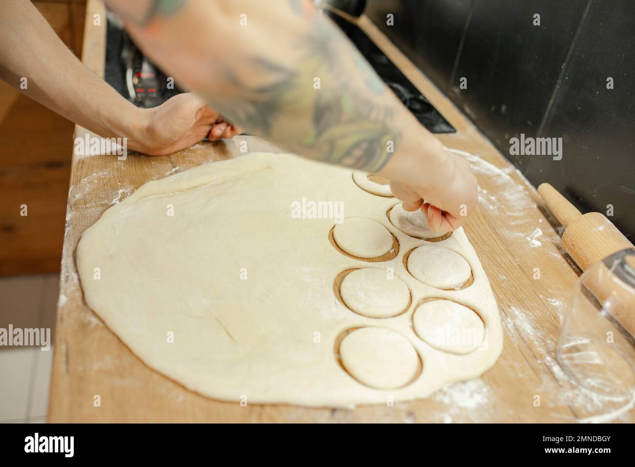 Male hands cut out round pieces with glass from raw thin dough. Roll out dough with rolling pin on kitchen tabletop. Stock Photo