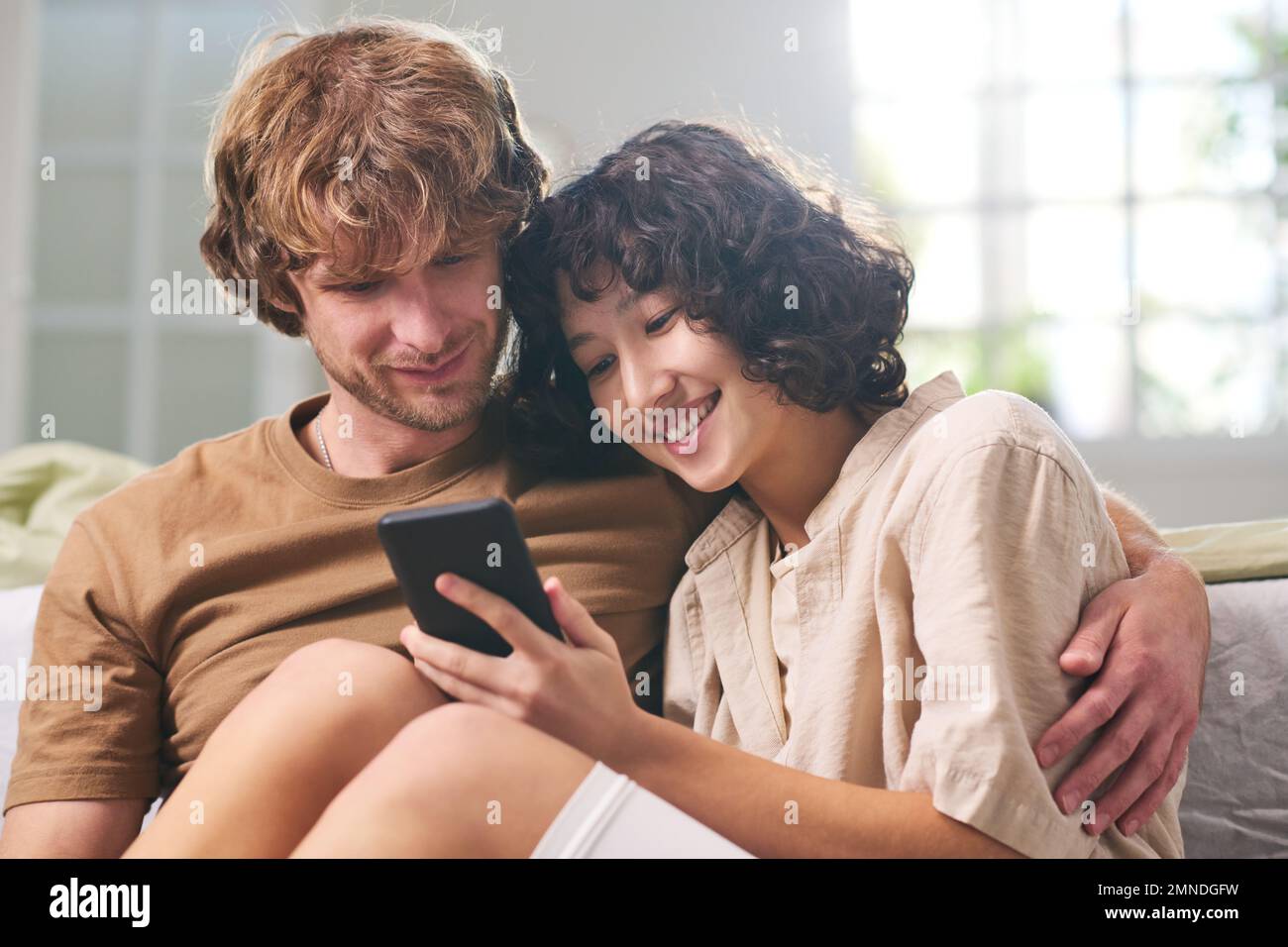 Young happy woman and her husband looking at mobile phone screen held by wife while relaxing at home and watching online video Stock Photo