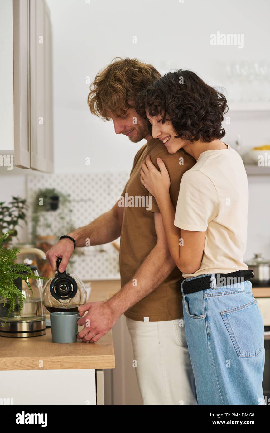 Side view of young woman embracing her husband with pot pouring tea or coffee in mug after preparing it for himself and his wife Stock Photo