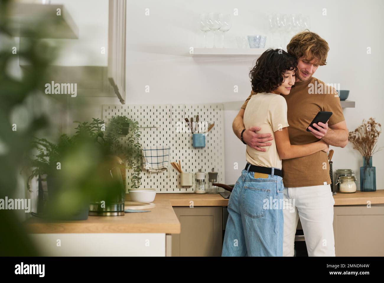 Young woman embracing her husband and both looking at screen of smartphone held by man while standing in the kitchen Stock Photo