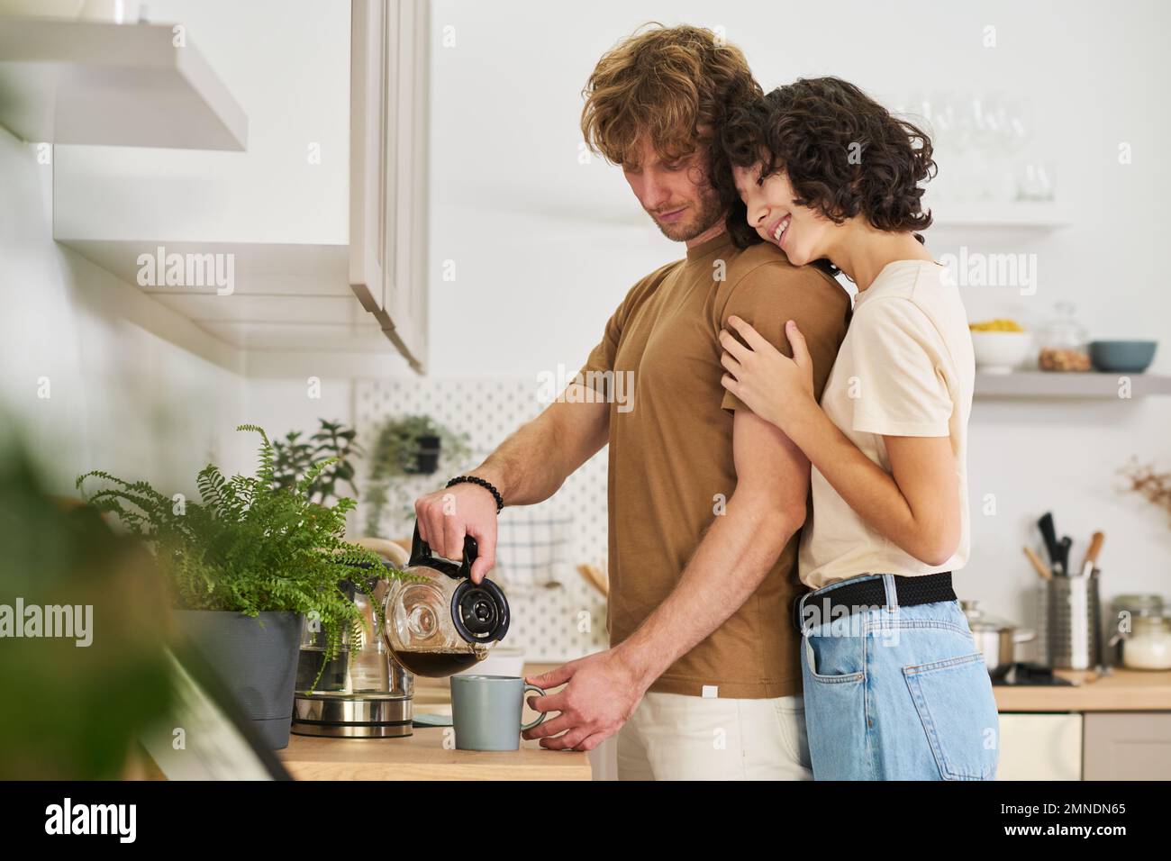 Young smiling woman standing close to her husband pouring coffee in grey mug and embracing him while looking forwards for breakfast Stock Photo