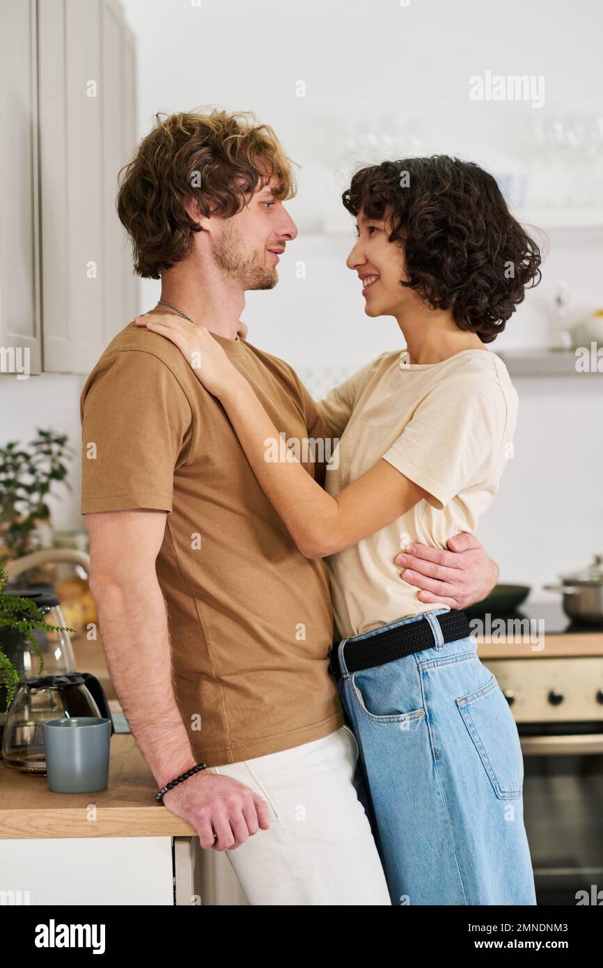 Happy young husband and wife in casualwear looking at one another while man embracing woman who keeping her hand on his shoulder Stock Photo