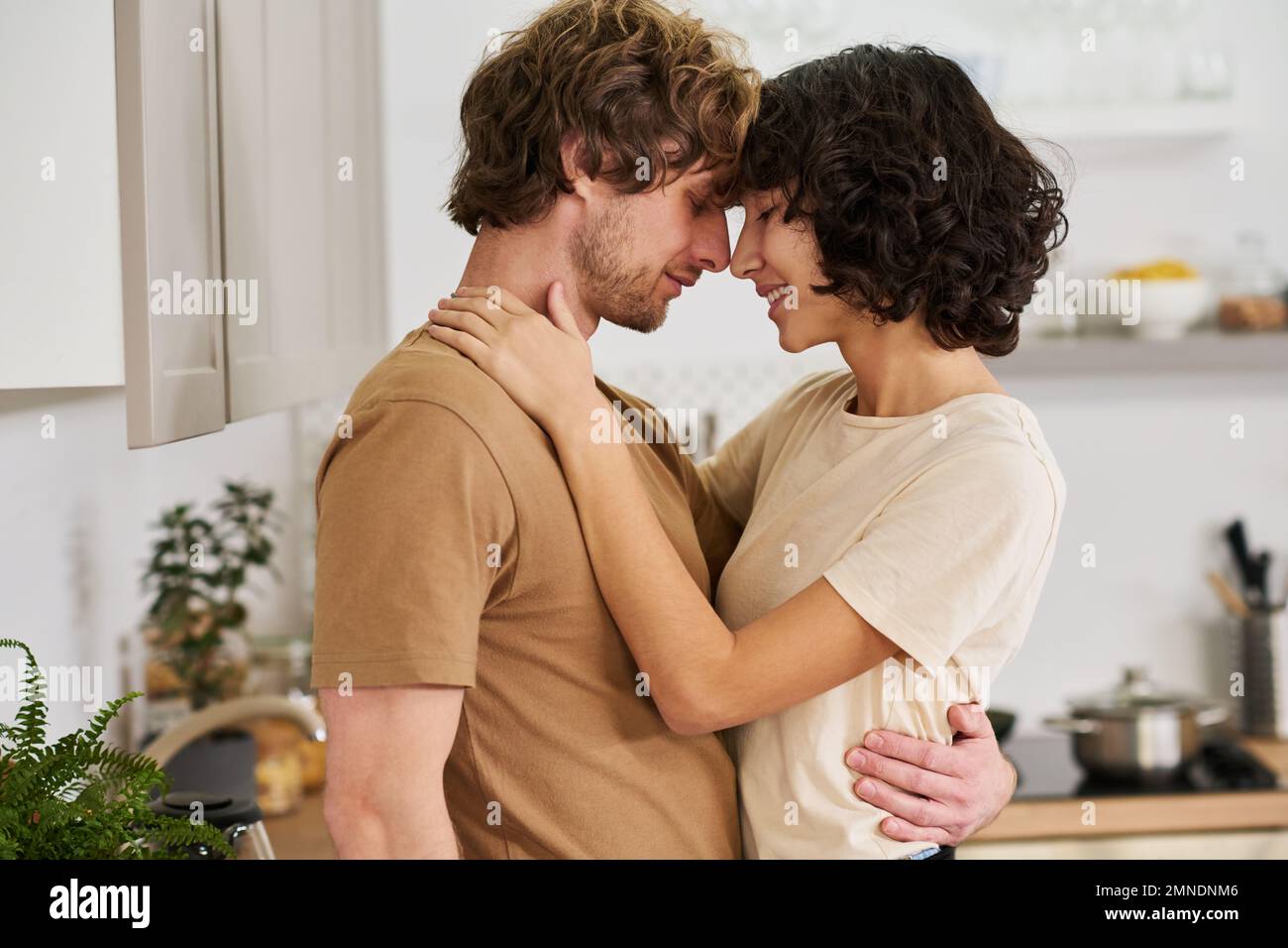 Side view of young amorous husband and wife touching by their foreheads and noses while woman keeping hands on shoulders of her husband Stock Photo