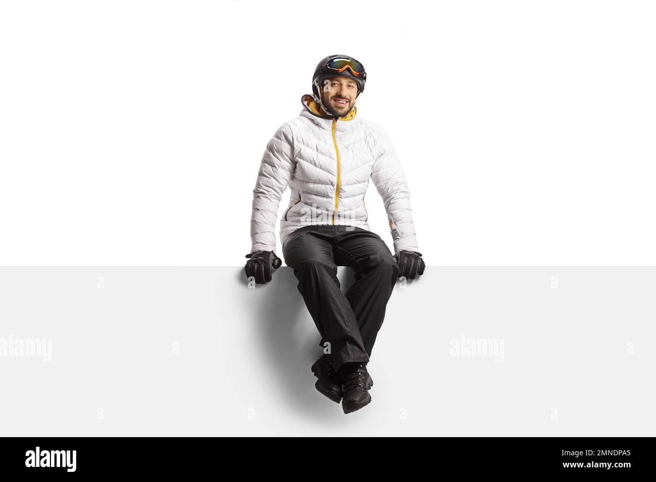 Young man wearing a skiing equipment and sitting on a blank panel isolated on white background Stock Photo