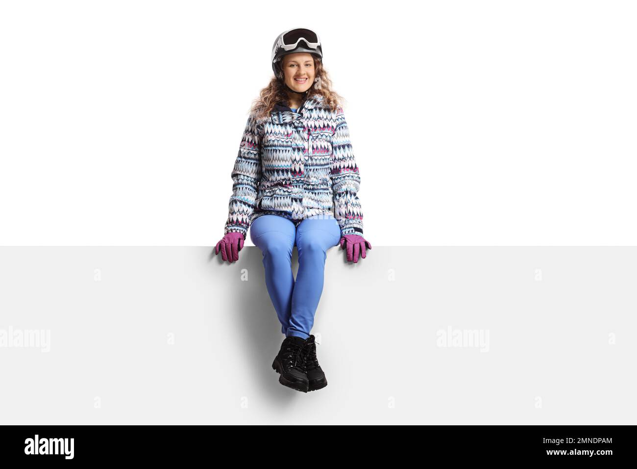 Young woman wearing a skiing equipment and sitting on a blank panel isolated on white background Stock Photo