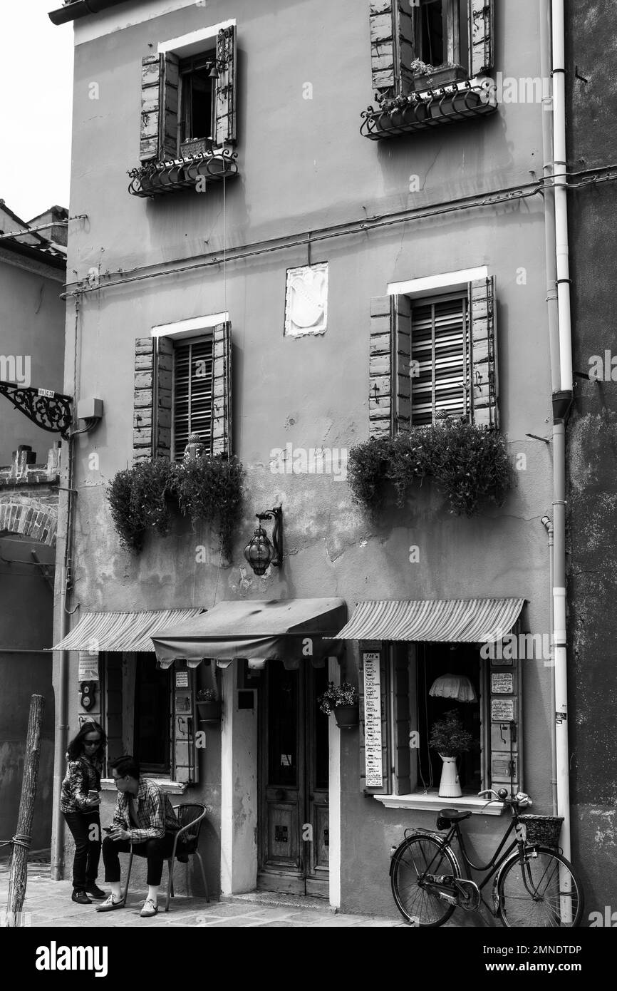 A Young man rests outside of a brightly painted shop on Burano, searching his phone, a bicycle is leant against the wall Stock Photo