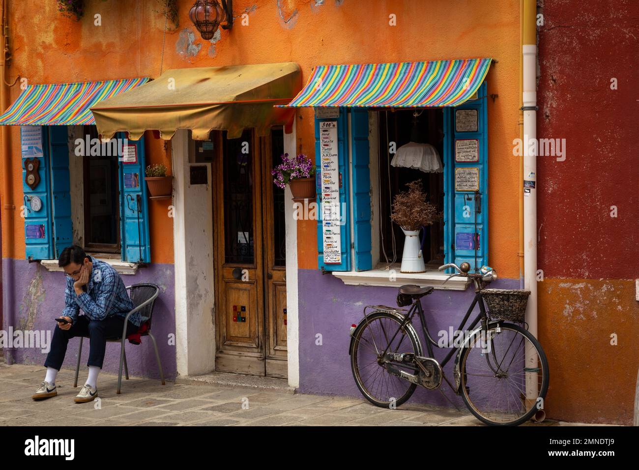 A Young man rests outside of a brightly painted shop on Burano, searching his phone, a bicycle is leant against the wall Stock Photo