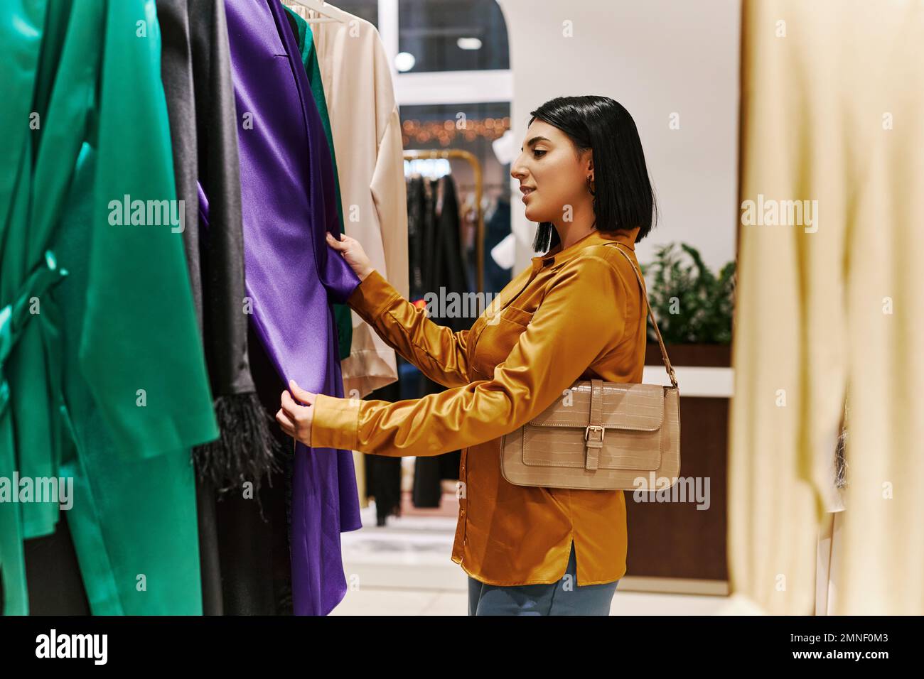 Side view portrait of smiling young woman browsing clothes on racks in luxury boutique and enjoying shopping Stock Photo