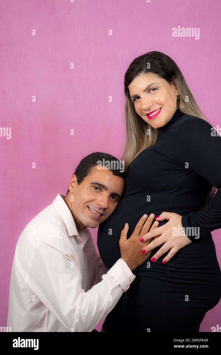 Husband listening to the baby inside his pregnant wifes belly. Isolated against pink background. Stock Photo
